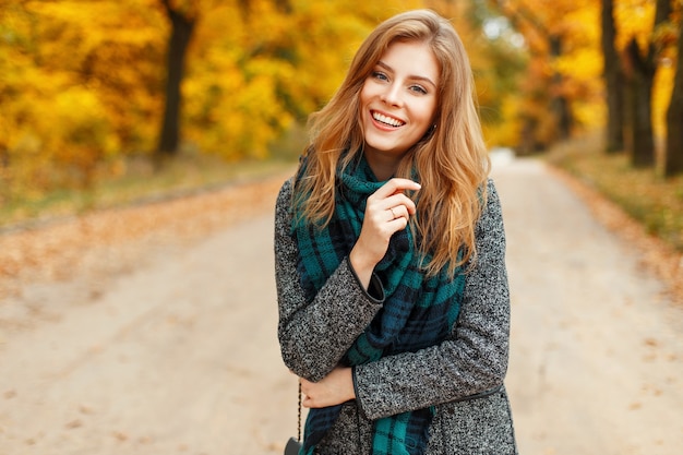 Glückliche schöne Frau mit einem Lächeln in einem trendigen Herbstmantel mit einem Schal auf einem Hintergrund des gelben Laubs