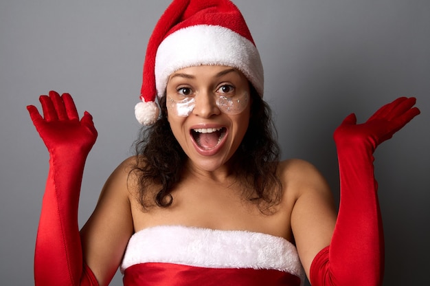 Glückliche schöne Frau mit glänzenden, glättenden Augenflecken unter den Augen, gekleidet in Santa-Kleidung, legt ihre Hände nach oben und freut sich beim Blick in die Kamera. Frohe Weihnachten, Neujahr und Kosmetikkonzept