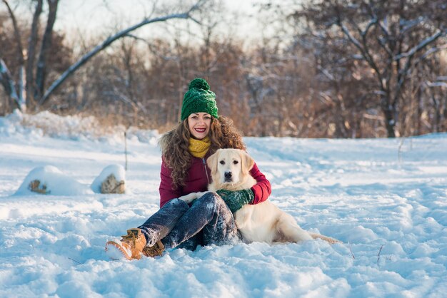 Glückliche schöne junge Frau, die Schneeflocken von ihren Händen zu ihrem Hund Golden Retriever an einem Wintertag bläst.