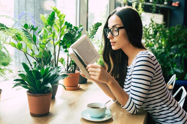 Glückliche schöne junge Frau in Gläsern liest Buch und trinkt Kaffee im modernen Café