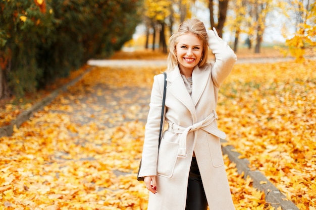 Glückliche schöne junge Frau mit einem Lächeln in einem trendigen klassischen Mantel mit einer Handtasche posiert Spaziergänge im Herbsttag in einem gelben Laub