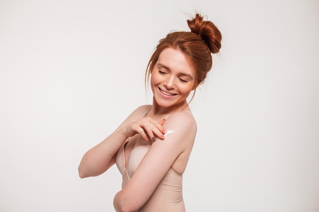 Glückliche schöne junge rothaarige frische Frau mit süßem Lächeln in beigem BH, die Creme auf ihrer Schulter im Studio auf weißem Hintergrund aufträgt Schönheit, Gesundheit und Hautpflege
