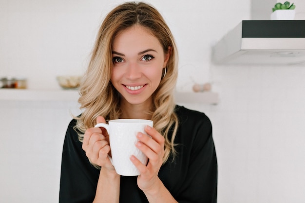 Glückliche schöne kaukasische blonde Frau in Nachtwäsche trinkt Kaffee in der Küche und lächelt in die Kamera zu Hause, wachte am Morgen an einem sonnigen Tag auf.