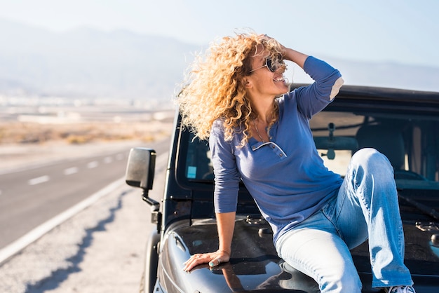 Glückliche schöne reife Frau mit Sonnenbrille, die mit der Hand im lockigen Haar posiert, während sie auf der Motorhaube des Jeeps auf der Autobahn sitzt. Fröhliche, stilvolle Frau, die während ihres Roadtrips auf der Motorhaube am Straßenrand posiert