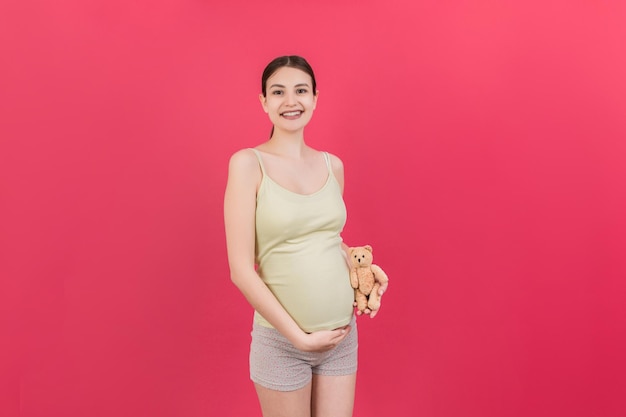 Glückliche schwangere Frau, die einen Teddybären gegen ihren Bauch auf farbigem Hintergrund hält Junge Mutter erwartet ein Baby Kopieren Sie Platz