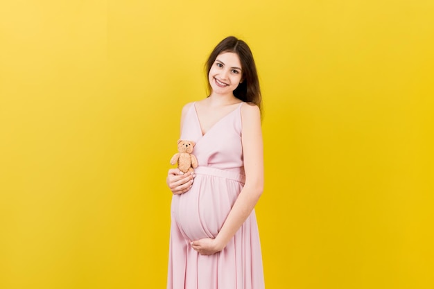 Glückliche schwangere Frau, die einen Teddybären gegen ihren Bauch auf farbigem Hintergrund hält Junge Mutter erwartet ein Baby Kopieren Sie Platz