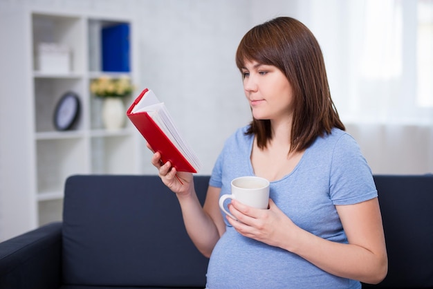 Glückliche schwangere Frau, die zu Hause ein Buch liest und Tee oder Kaffee trinkt