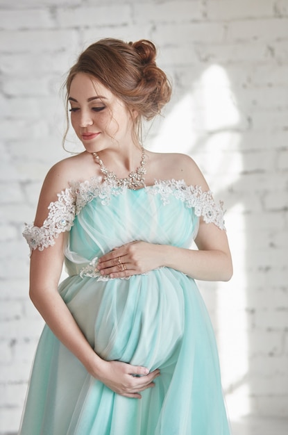 Glückliche schwangere Frau im langen Abendkleid, die Bauchhände berührt. Warten auf die Geburt eines Kindes, eine Frau im achten Monat der Schwangerschaft. Luxuriöses schwangeres Mädchen in einem azurblauen Kleid