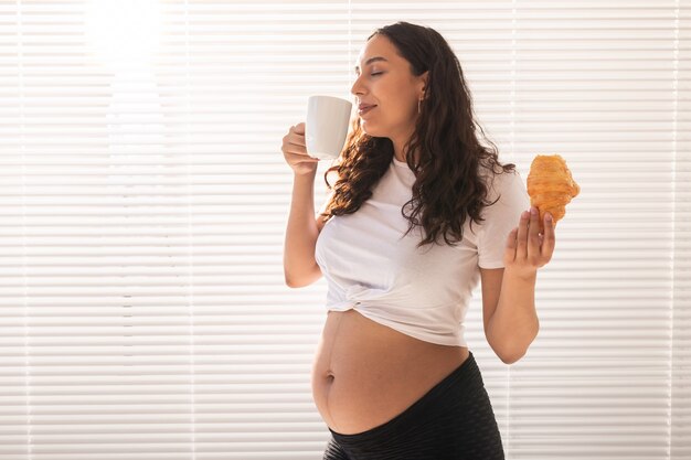 Glückliche schwangere frau mit croissantbrötchen zu hause, schwangerschaftsessen und menschenkonzept