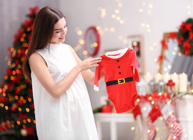 Glückliche schwangere Frau mit Weihnachtsmann-Babyanzug im weihnachtlich dekorierten Raum