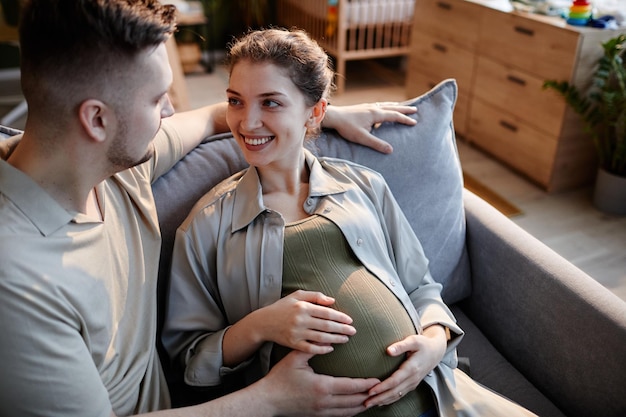 Glückliche schwangere Frau sitzt mit ihrem Mann auf dem Sofa und lächelt, während er ihren Bauch streichelt