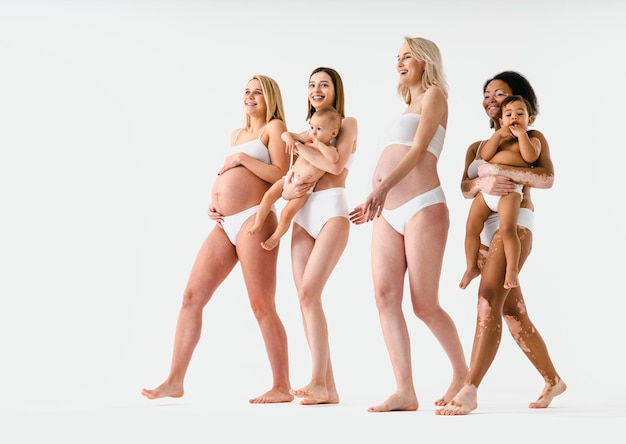 Foto glückliche schwangere frauen mit dickem bauch und schöne mütter mit babys im studio - multirassische gruppe schwangerer frauen und mütter mit kindern in unterwäsche - schwangerschaft, mutterschaft, menschen, körperhaltung