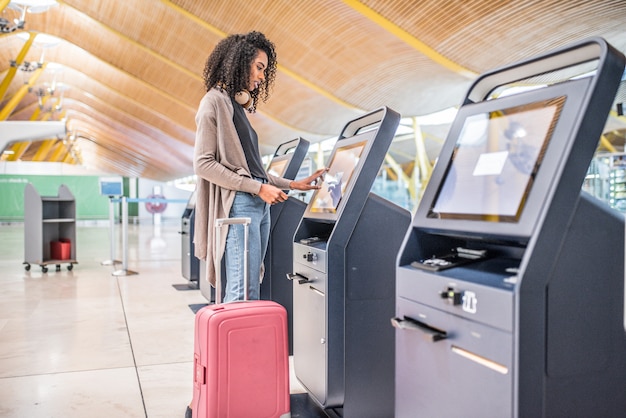 Glückliche schwarze Frau, die den Abfertigungsautomaten am Flughafen erhält die Bordkarte verwendet.
