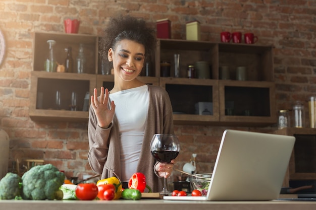 Glückliche schwarze Frau, die Laptop benutzt und Wein im modernen Kücheninnenraum trinkt