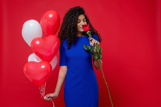 Glückliche schwarze Frau mit Herzen formte die Ballone und Rose, die auf roter Wand lokalisiert wurden