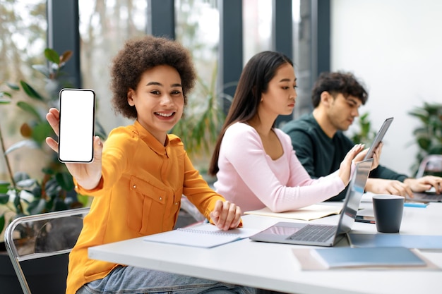 Glückliche schwarze Studentin, die im Klassenzimmer sitzt und ihr Telefon mit leerem Bildschirm für den Unterricht zeigt