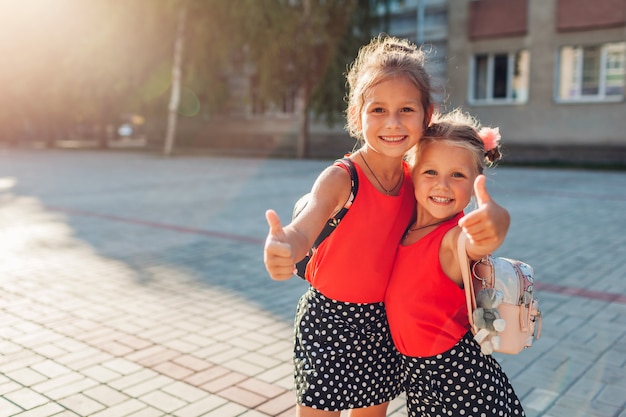 Glückliche Schwestermädchen, die Rucksäcke tragen und sich Daumen zeigen. Scherzt die Schüler, die draußen Schulgebäude lächeln. Bildung