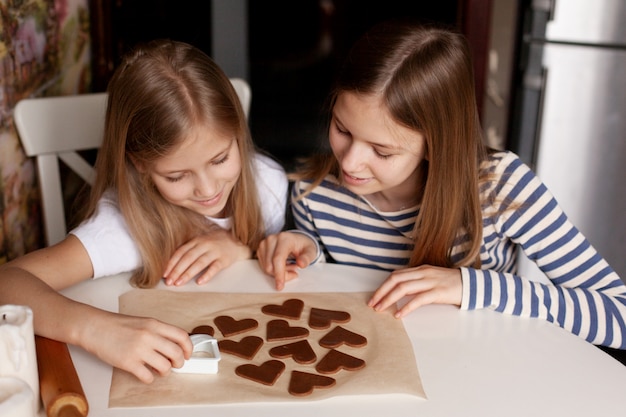 Glückliche Schwestern in der Wohnküche am Tisch schnitten herzförmige Kekse aus dem Teig