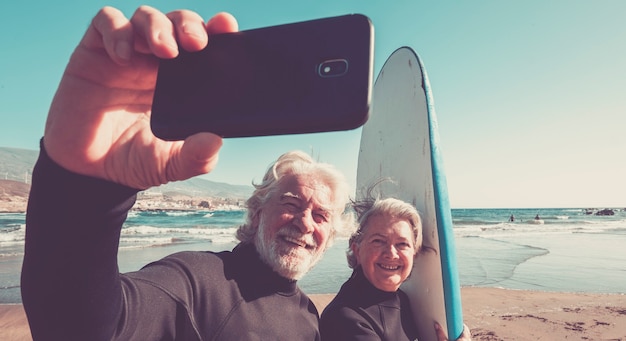 Glückliche Seniorenpaare am Strand, die versuchen zu surfen und zusammen Spaß zu haben - reife Frau und Mann heirateten und machen ein Selfie mit den Neoprenanzügen und Surftischen mit Meer oder Ozean im Hintergrund