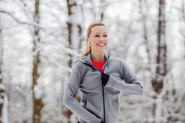 Glückliche Sportlerin, die Sweatshirt beim Stehen in der Natur am verschneiten Wintertag bindet.