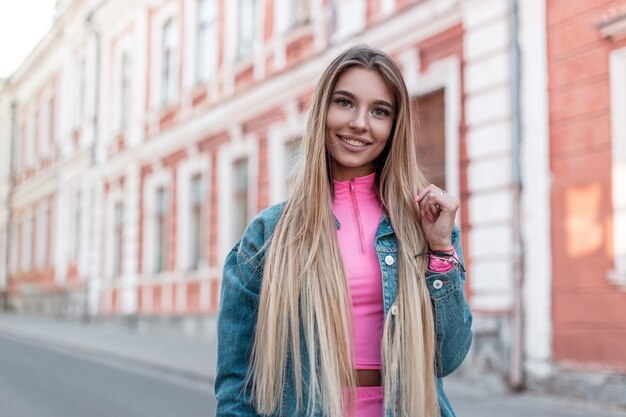 Glückliche städtische junge Frau mit positivem Lächeln mit blondem langem Haar in einer trendigen Jeansjacke in einem stilvollen rosa Oberteil stellt nahe einem weißen Weinlesegebäude in der Straße auf. Ziemlich fröhliches Mädchen geht in die Stadt.