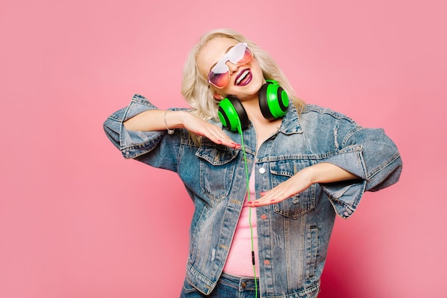 Glückliche stilvolle blonde Frau in rosa Brille mit großen Kopfhörern, die Musik hören und auf farbigem Hintergrund tanzen