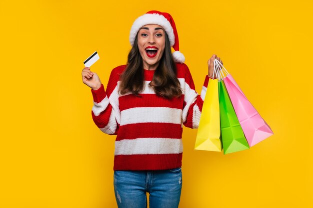 Glückliche stilvolle schöne Frau in Weihnachtsmütze mit bunten Einkaufstüten isoliert auf gelbem Hintergrund