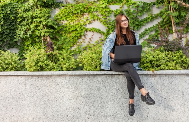 Glückliche Studentin in Jeansjacke und Brille benutzt Laptop, während sie im Stadtpark sitzt