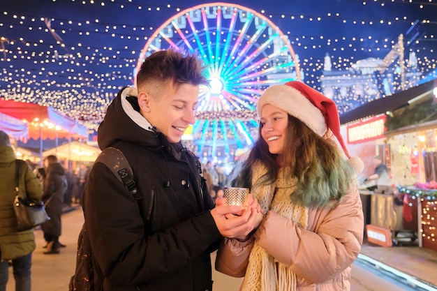 Glückliche Teenager paaren Jungen und Mädchen in Weihnachtsmütze auf dem Weihnachtsmarkt, die Hände mit Kerze wärmen. Lichter, helle Girlanden der Abendstadt, Riesenradhintergrund