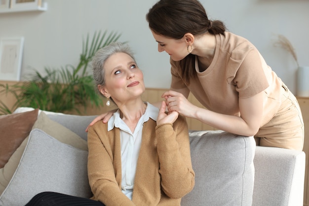 Glückliche Tochter, die ältere Mutter umarmt, hinter dem Sofa im Wohnzimmer steht und den zarten Moment zu Hause genießt