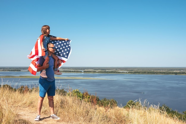 Glückliche Tochter mit amerikanischer Flagge, die auf den Schultern der Väter sitzt, genießt die Natur