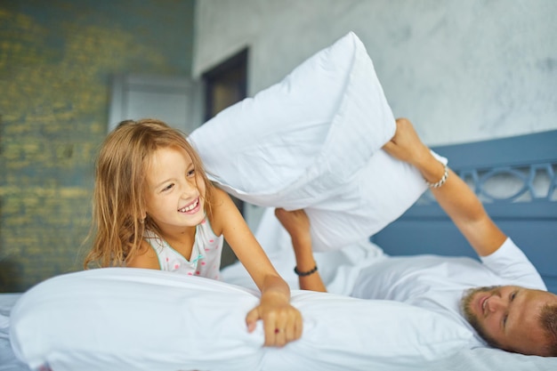 Glückliche Tochter und Vater spielen morgens zu Hause Kissenschlacht im Bett, kämpfen mit Kissen, haben Spaß beim gemeinsamen Spielen zu Hause am Familienwochenende