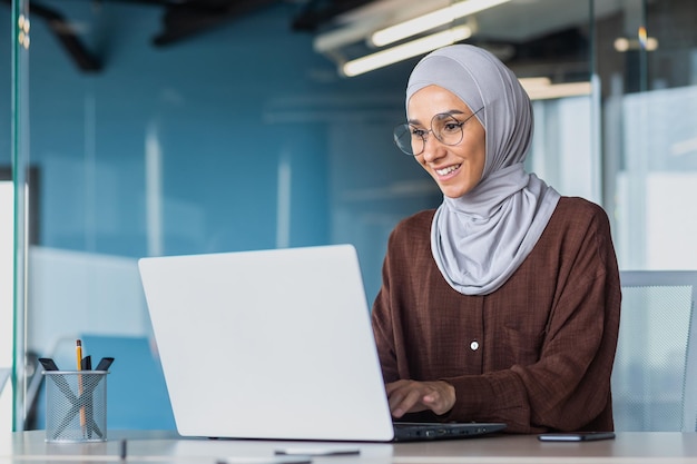 Glückliche und erfolgreiche Geschäftsfrau im Hijab, die mit Laptop in einem modernen Bürogebäude arbeitet