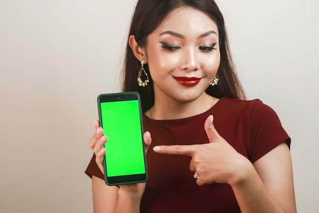 Glückliche und lächelnde junge asiatische Frau, die auf einen grünen leeren Bildschirm zeigt und zeigt