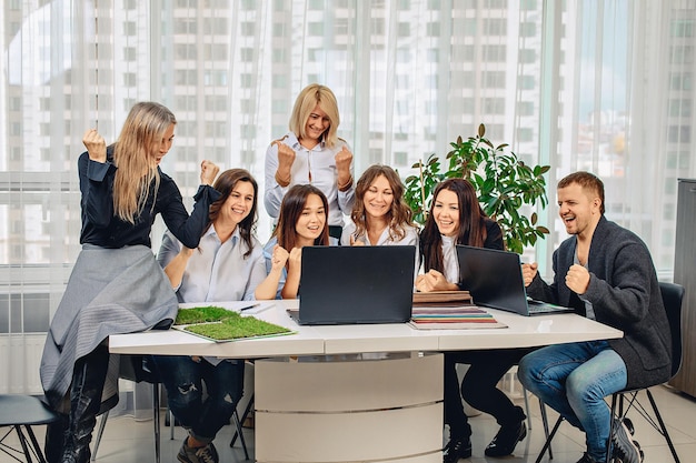 Glückliche und schöne Mitarbeiter, die die gute Nachricht erfahren haben, sitzen am selben Tisch und lächeln im Büro. Lustiges Konzept