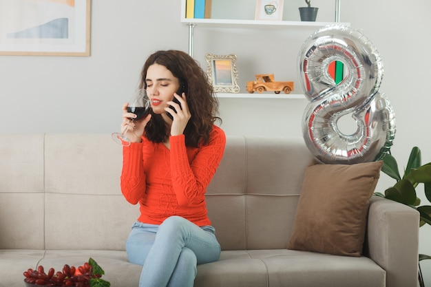 Glückliche und zufriedene junge Frau, die fröhlich auf einer Couch sitzt und mit einem Glas Wein im hellen Wohnzimmer telefoniert und den internationalen Frauentag am 8. März feiert