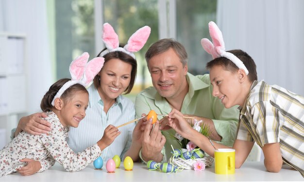 Glückliche vierköpfige Familie, die Hasenohren trägt und zu Hause Ostereier malt