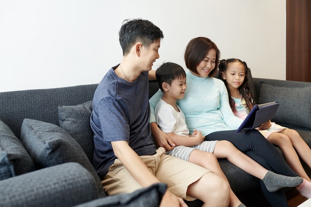 Glückliche vietnamesische Familie mit zwei Kindern, die es genießen, Zeit zusammen zu verbringen und animierte Videos oder Blogs auf einem digitalen Tablet anzusehen