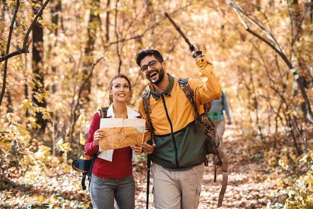 Glückliche Wanderer, die Wald im Herbst erkunden. Frau, die Karte hält, während Mann mit Stock auf den richtigen Weg zeigt.
