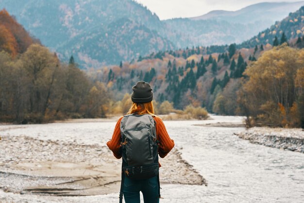 Glückliche Wanderin mit Rucksack am Flussufer blickt in die Berge und die herbstliche Waldnatur. Hochwertiges Foto