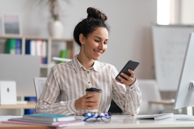 Glückliche weibliche Angestellte, die Smartphone benutzt und Kaffee im Büro trinkt und eine Pause bei der Arbeit hat