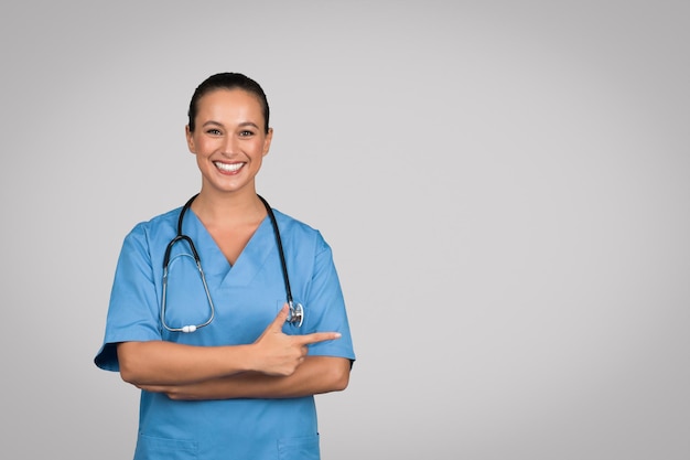 Glückliche weibliche Ärztin oder Krankenschwester in blauer Uniform, die auf einen Kopierplatz auf grauem Hintergrund zeigt