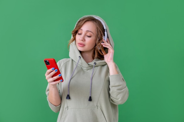 Glückliche Zähne Lächeln Frau hören Musik Kopfhörer mit Smartphone in der Hand suchen Bildschirm Kaukasische Frau genießen Podcast oder Hörbücher gekleidet übergroßen Hoodie grünen Hintergrund Nahaufnahme Porträt