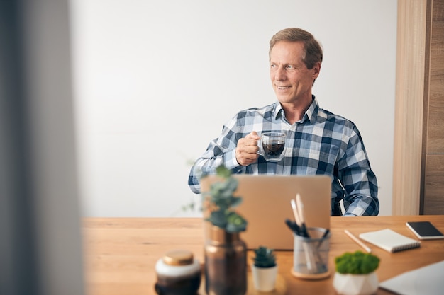 Glückliche, zufriedene erwachsene männliche Person, die eine Tasse in der rechten Hand hält und Kaffee trinken geht, während sie mit dem Laptop arbeitet