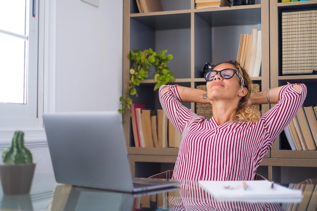 Glückliche zufriedene kaukasische Frau, die sich zu Hause im Büro ausruht, sitzt mit Laptop, hält die Hände hinter dem Kopf