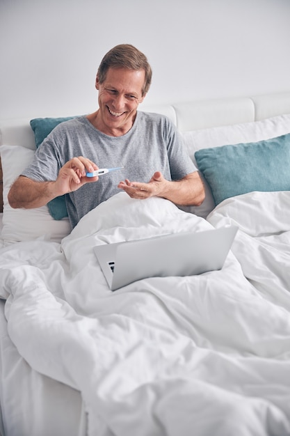 Glückliche, zufriedene männliche Person, die ein Lächeln auf dem Gesicht behält, während sie eine medizinische Online-Beratung im Schlafzimmer hat