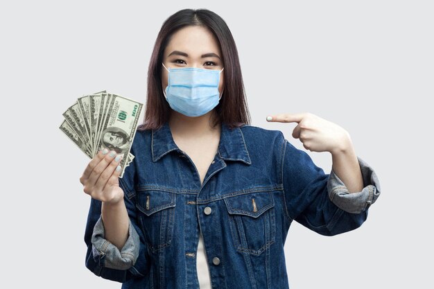 Glückliche zufriedene schöne junge Geschäftsfrau mit medizinischer Maske in blauer Jeansjacke, die viele Dollar hält, mit dem Finger zeigt und in die Kamera schaut. Innen, Studioaufnahme, isoliert, grauer Hintergrund