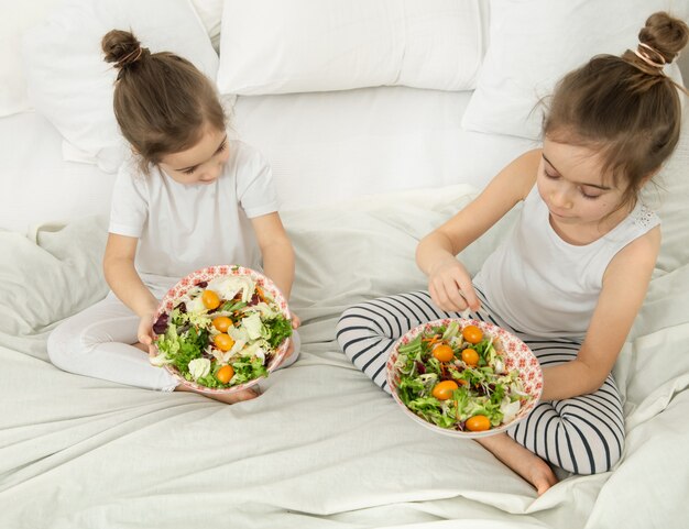 Glückliche zwei niedliche Kinder, die Gemüsesalat im Schlafzimmer auf dem Bett essen. Gesundes Essen für Kinder und Jugendliche.