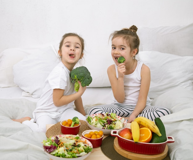 Glückliche zwei niedliche Mädchen, die Obst und Gemüse im Schlafzimmer auf dem Bett essen. Gesundes Essen für Kinder und Jugendliche.