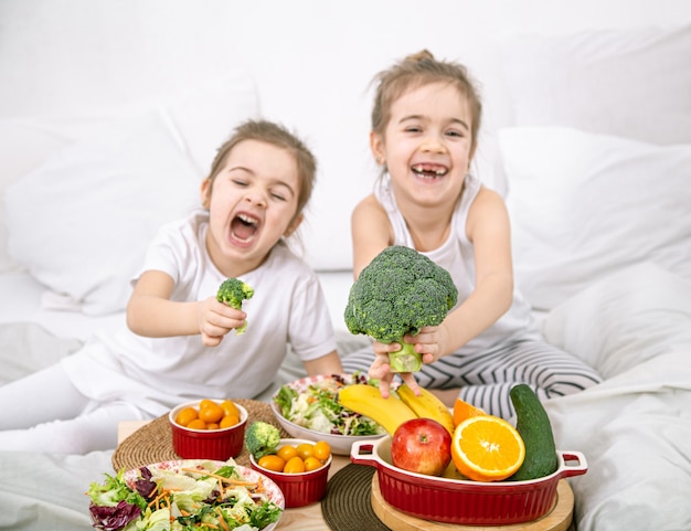 Glückliche zwei niedliche Mädchen, die Obst und Gemüse im Schlafzimmer auf dem Bett essen. Gesundes Essen für Kinder und Jugendliche.